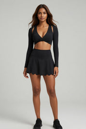 A woman modeling a black two-piece athletic set consisting of a long-sleeve crop top with side cutouts and a matching flared mini skirt with a scalloped edge, paired with black sneakers, against a light gray studio background.