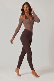 A woman modeling a two-piece activewear set consisting of a long-sleeve scoop-neck bodysuit in taupe and matching high-waisted leggings in dark brown, set against a plain light gray studio background.