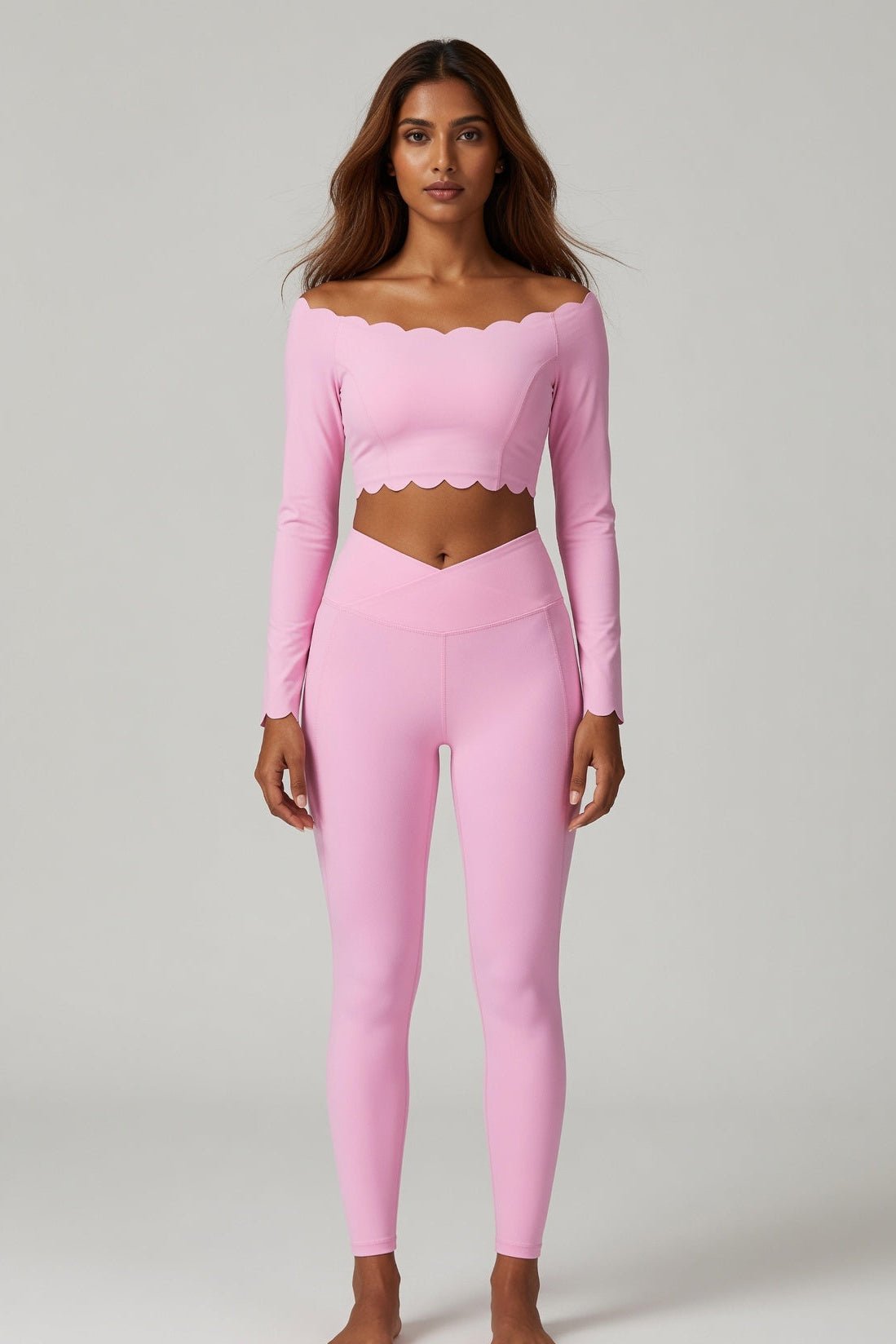 A woman modeling a light pink two-piece activewear set consisting of a long-sleeve cropped top with a scalloped neckline and matching high-waisted leggings, shot against a neutral light gray background.
